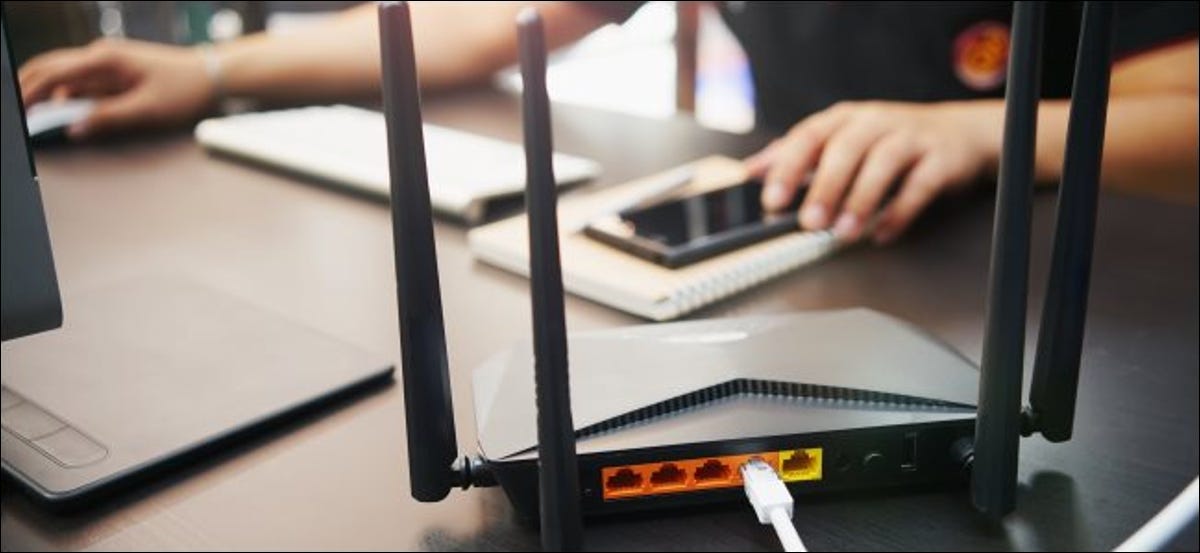 Un enrutador inalámbrico sobre una mesa con un hombre que usa una computadora de escritorio Mac y un iPhone en el fondo.